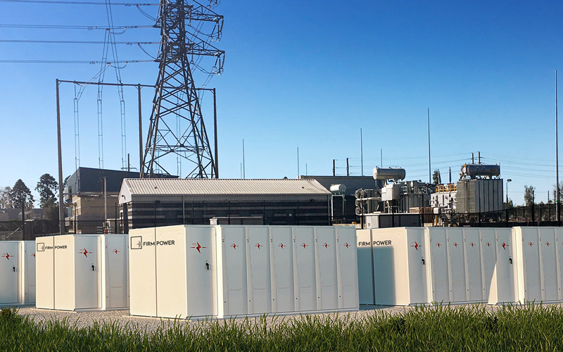 Battery Energy Storage System buildings set up on some land with electricity lines and transmission line in background.